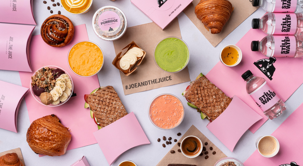 Assorterte Joe & The Juice-produkter, inkludert smørbrød, croissanter, grøt, juicer, açaí-boller og kaffe på en hvit bakgrunn med rosa emballasje og flasker med stille vann