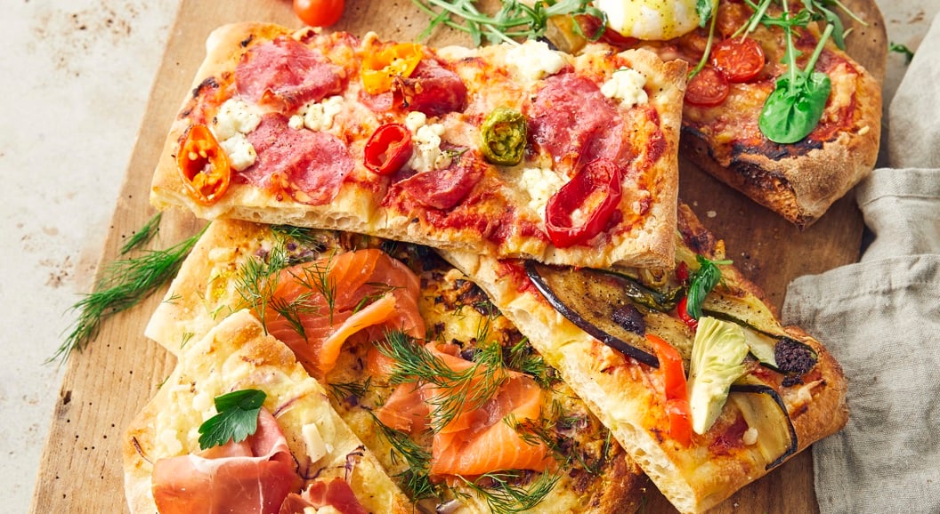 Assorterte gourmetflatbrødpizzaer på et trefat med toppings som inkluderer prosciutto, røkt laks, ruccola og ferske tomater, ledsaget av dippesauser på siden