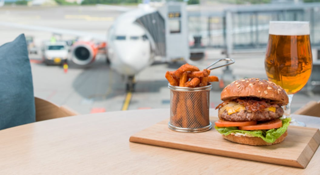 Gourmetburger med pommes frites og et glass øl på et bord med utsikt over en rullebane på flyplassen med et parkert fly i bakgrunnen