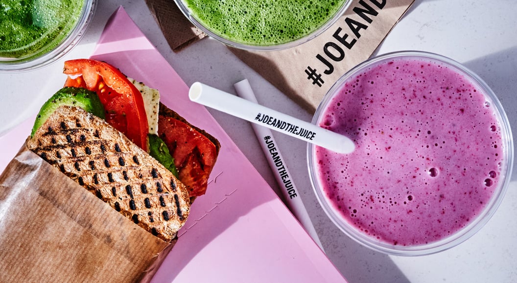 Juice bar-måltid med en grillet sandwich med avokado og tomat, ved siden av grønne og lilla smoothies på et bord, merket med #JoeAndTheJuice