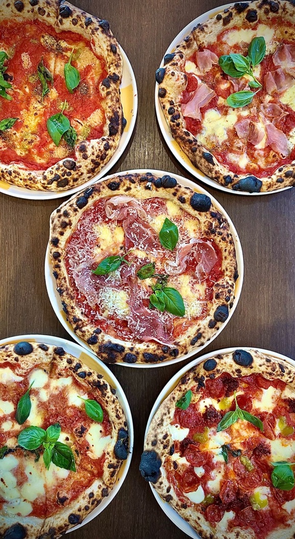 Fem håndverks-pizzaer med ulike toppinger inkludert basilikum, ost og spekemat på et trebord