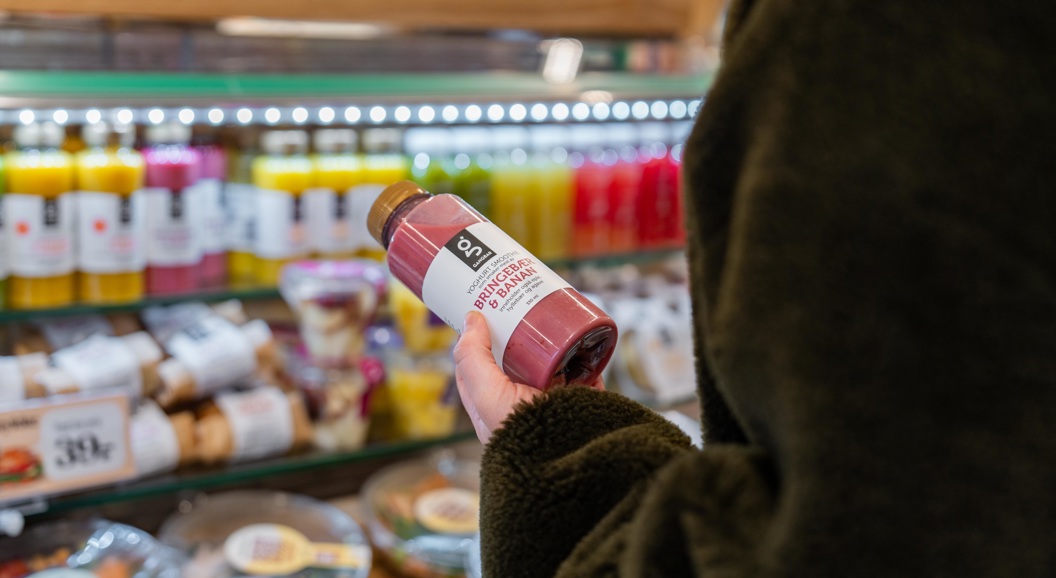 Person holder en flaske rosa smoothie i en butikk, foran en kjøledisk med ulike juicer og drikker