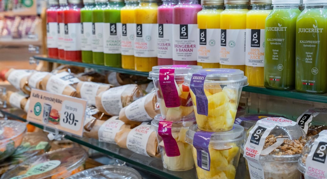 Supermarket shelf displaying assorted fresh juices in colorful bottles, deli wraps, and snack containers with pineapple chunks and granola yogurt