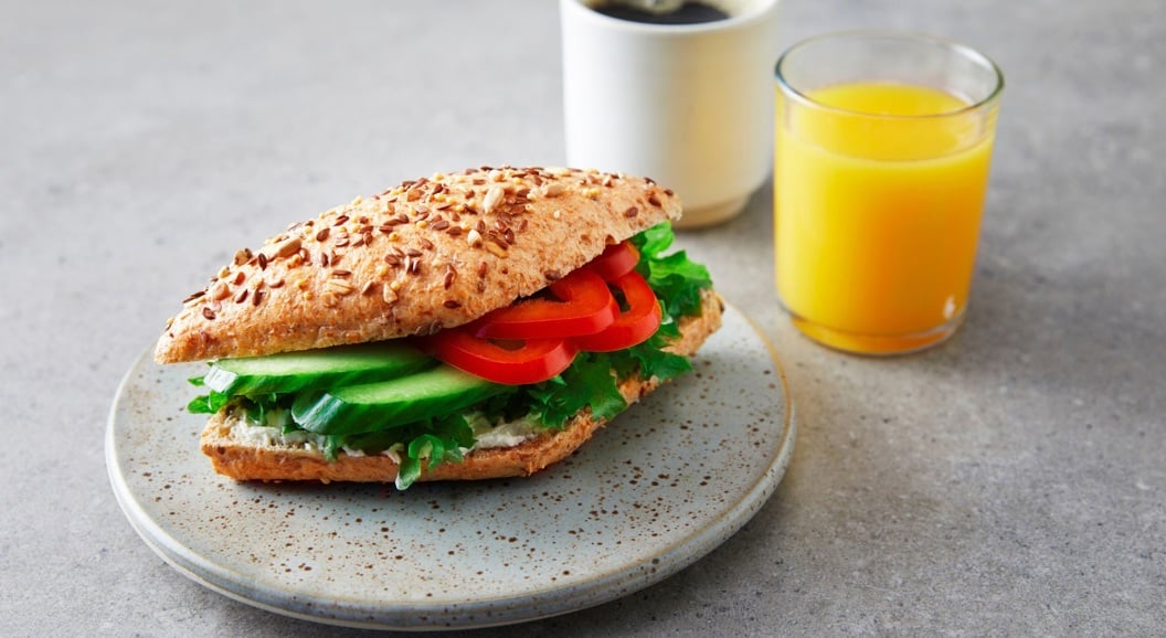 Frokostsandwich med egg, salat og tomat, servert med kaffe i pappkop og et glass appelsinjuice på trebrett