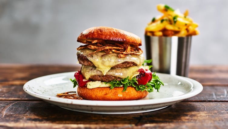Gourmet cheeseburger med sprøstekt løk, salat og tomater på en ristet bolle, servert med en side av gyldne pommes frites i en metallkopp