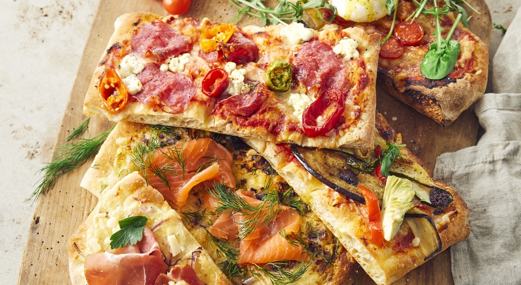 Assorted gourmet flatbread pizzas on a wooden board with toppings including prosciutto, smoked salmon, arugula, and fresh tomatoes, accompanied by dipping sauces on the side