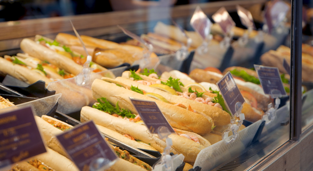 Freshly made baguette sandwiches displayed in a deli case, featuring a variety of fillings such as shrimp and greens, with price tags visible in the foreground