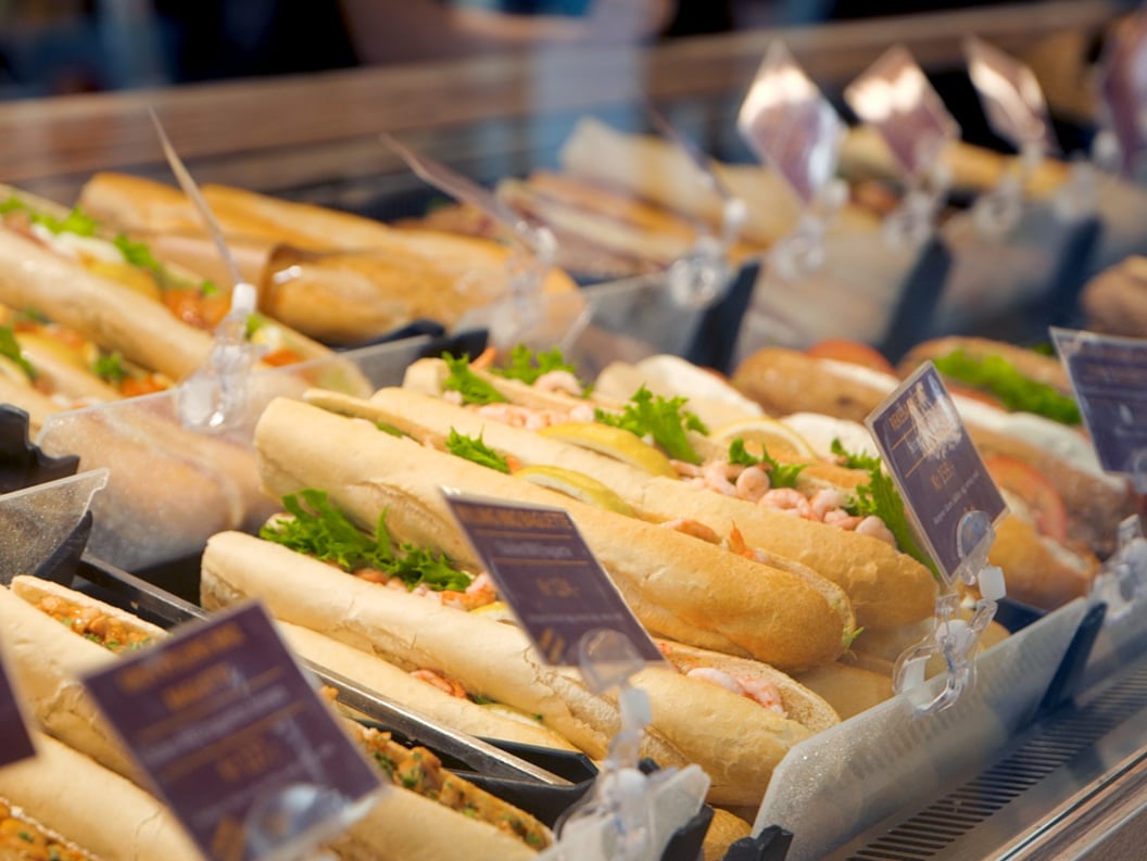 Nystekte baguettesmørbrød vist i en delikatedisk, med ulike fyllinger som reker og grønnsaker, med prislapper synlige i forgrunnen