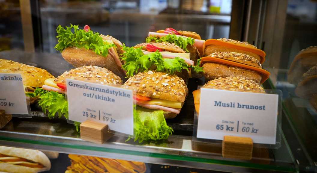 Assorted sandwiches with lettuce, cheese, and meats in a bakery display, featuring grain-topped buns with visible price tags in Norwegian krone