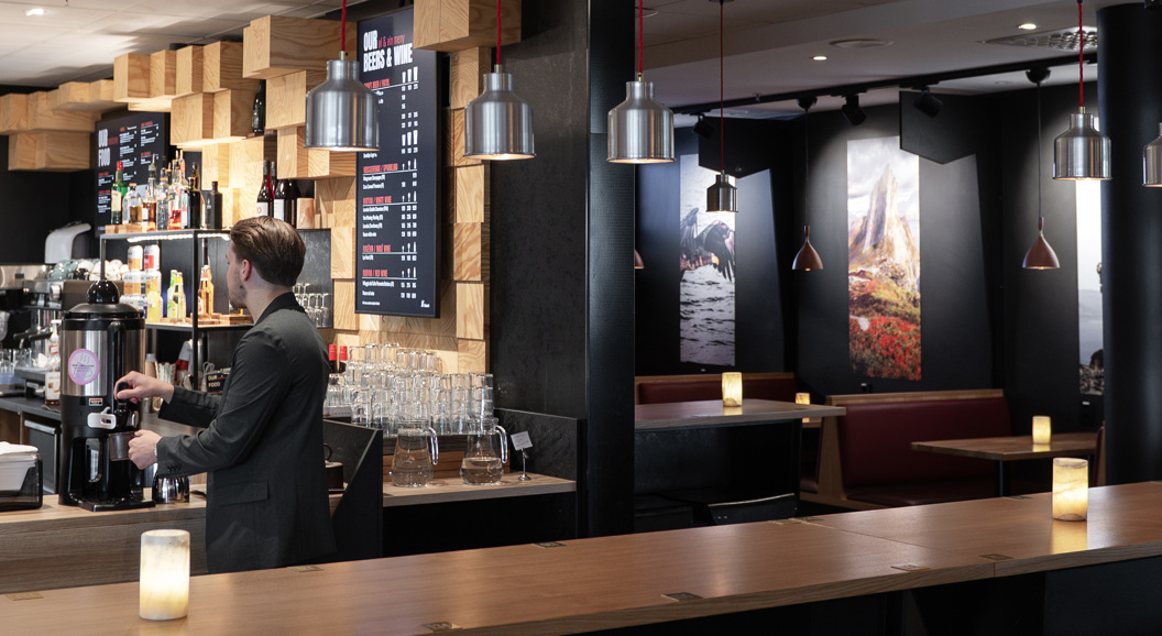 Koselig moderne kaféinteriør med en barista som tilbereder kaffe bak disken. Lokalet har moderne dekor, møbler i tre, hengende pendellamper, og innrammede landskapsbilder på veggene