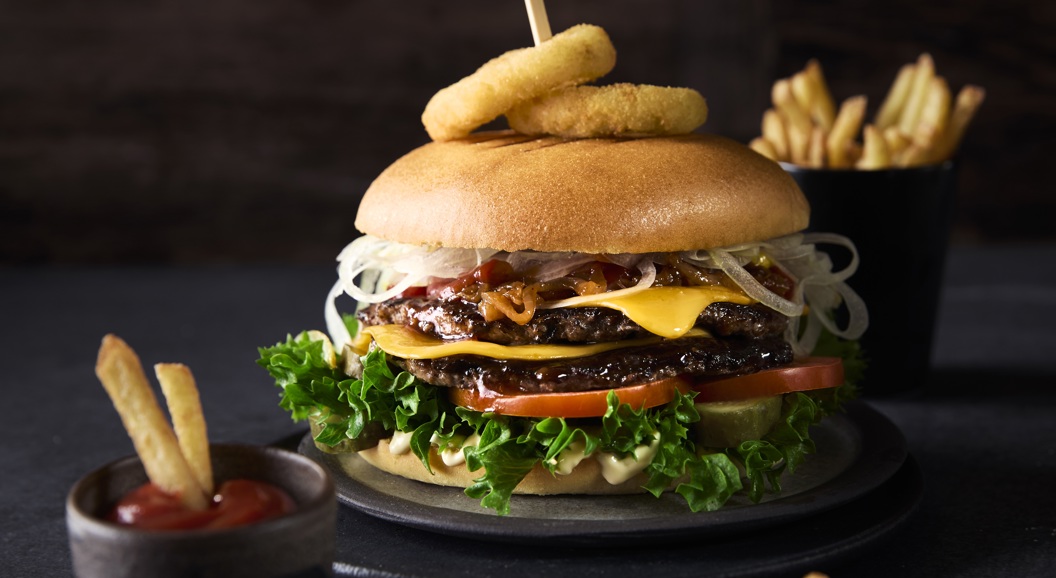 Delicious gourmet burger with lettuce, tomatoes, cheese, and onion rings on a bun, garnished with a skewer, accompanied by a side of fries and ketchup