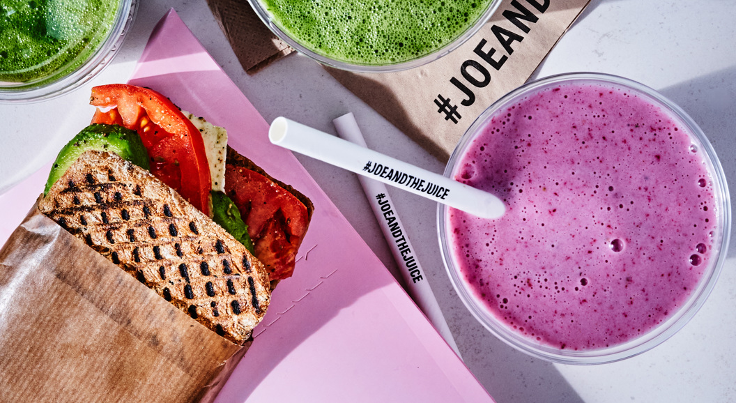 Juice bar meal featuring a grilled sandwich with avocado and tomato, alongside green and purple smoothies on a table, branded with #JoeAndTheJuice
