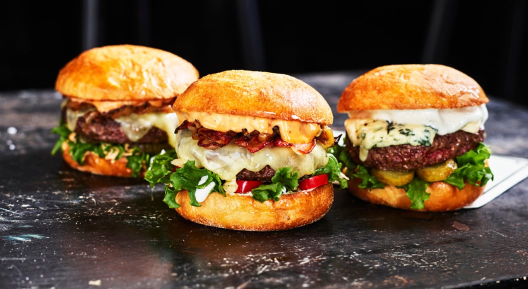 Tre gourmetburgere med ulike toppinger, inkludert salat, ost, pickles og bacon, på et rustikt svart bord