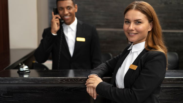 Hotellresepsjonens ansatte smiler, med en mann som svarer på telefonen og en kvinne som står i forgrunnen, begge iført formelle uniformer og navneskilt