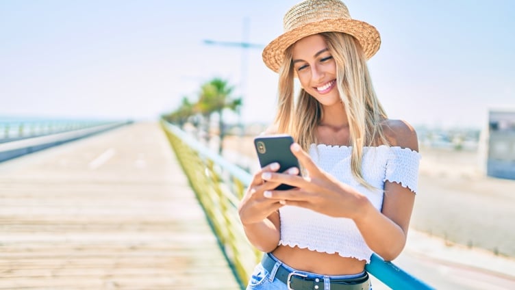 Kvinne i sommerantrekk og stråhatt smiler mens hun ser på telefonen sin, står på en promenade med palmer i bakgrunnen.
