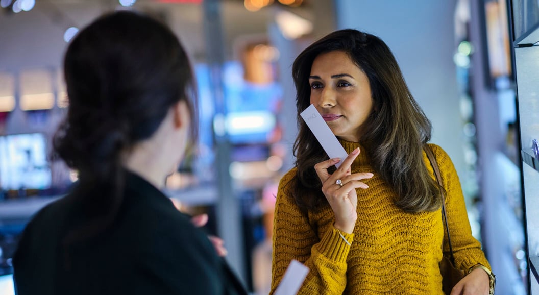 Kvinne i gul genser tester parfyme i en butikk, holder en duftstripe og snakker med en ansatt