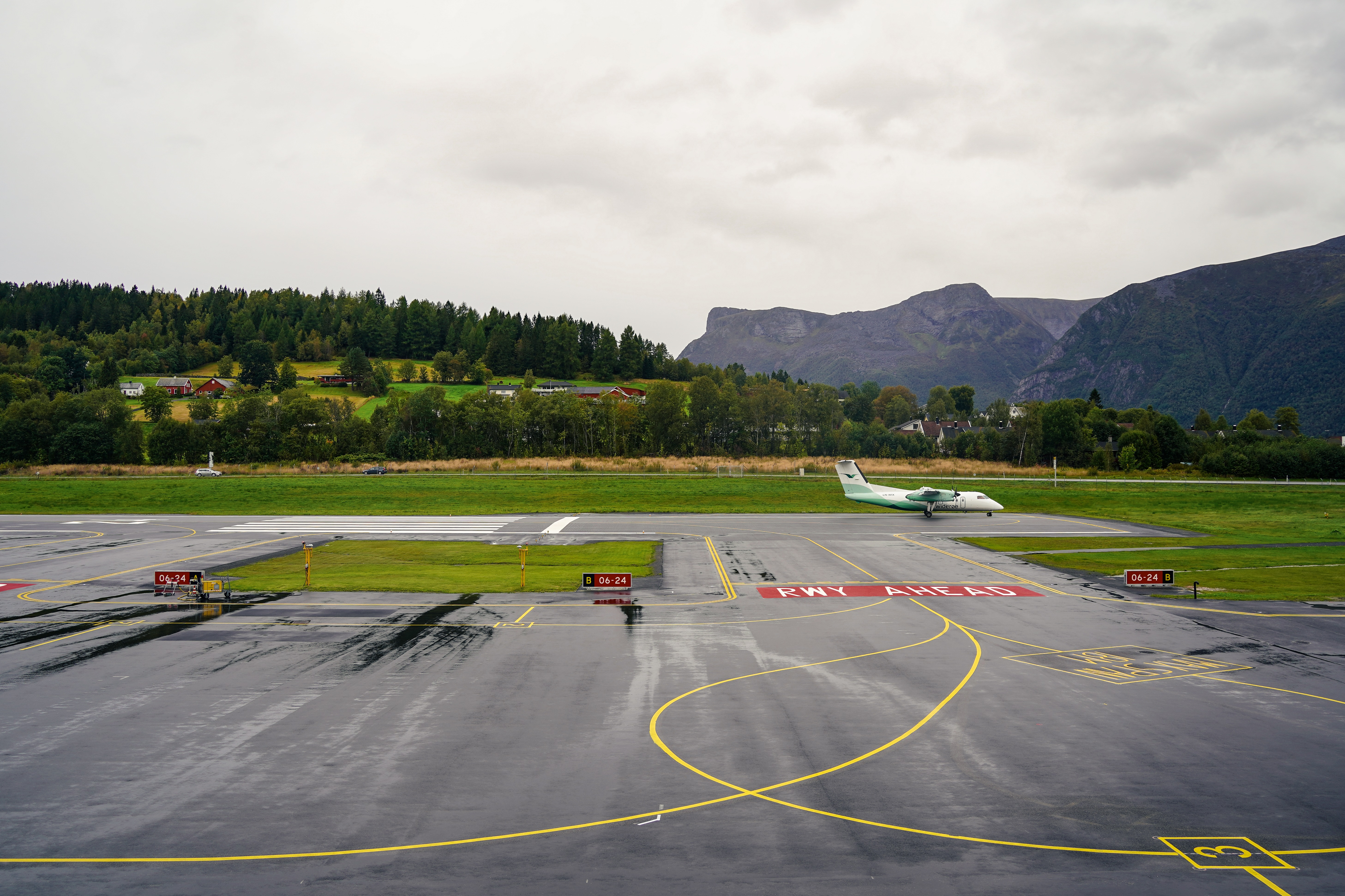 Flyplasslandskap med et lite passasjerfly på rullebanen, omgitt av grønne åser og fjell i bakgrunnen under en overskyet himmel.