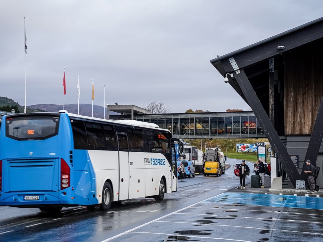 Buss ved terminal med reisende i høstvær, skilt mot taxiholdeplass i bakgrunnen.
