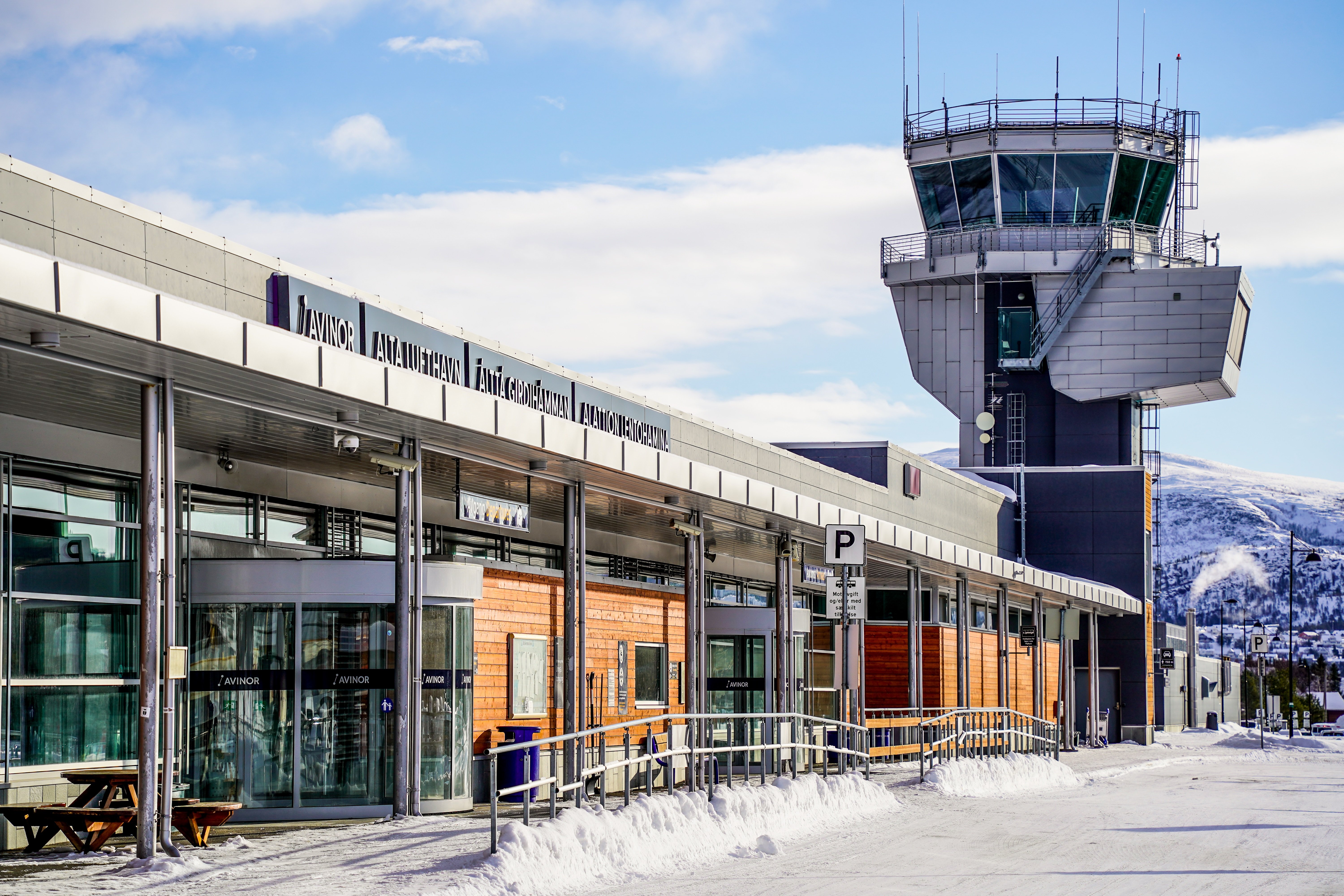 Flyplass med kontrolltårn i vinterlandskap, med skilt for Avinor Alta lufthavn, snødekket bakke og klare blå himmel.