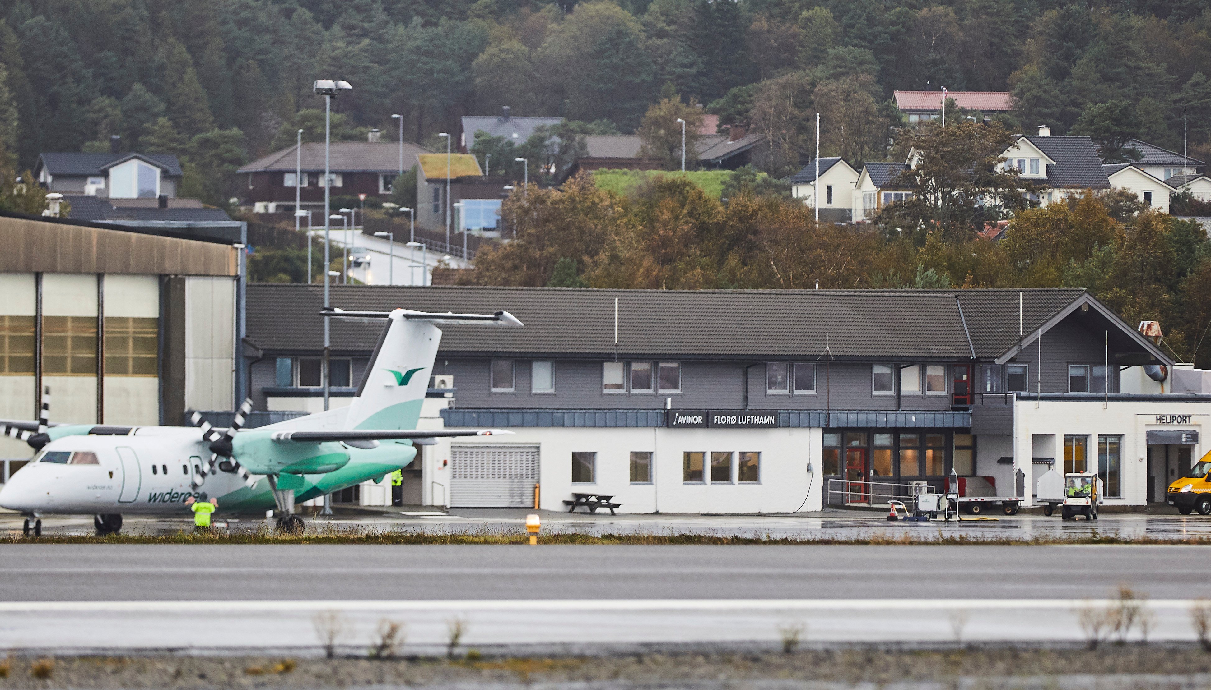 Widerøe-fly ved Florø lufthavn foran terminalbygningen, omgitt av skog og boliger i bakgrunnen.