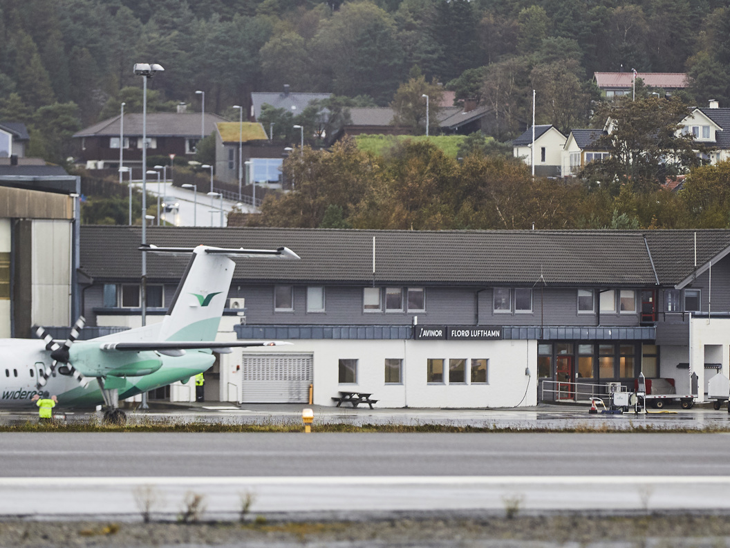 Widerøe-fly ved Florø lufthavn foran terminalbygningen, omgitt av skog og boliger i bakgrunnen.