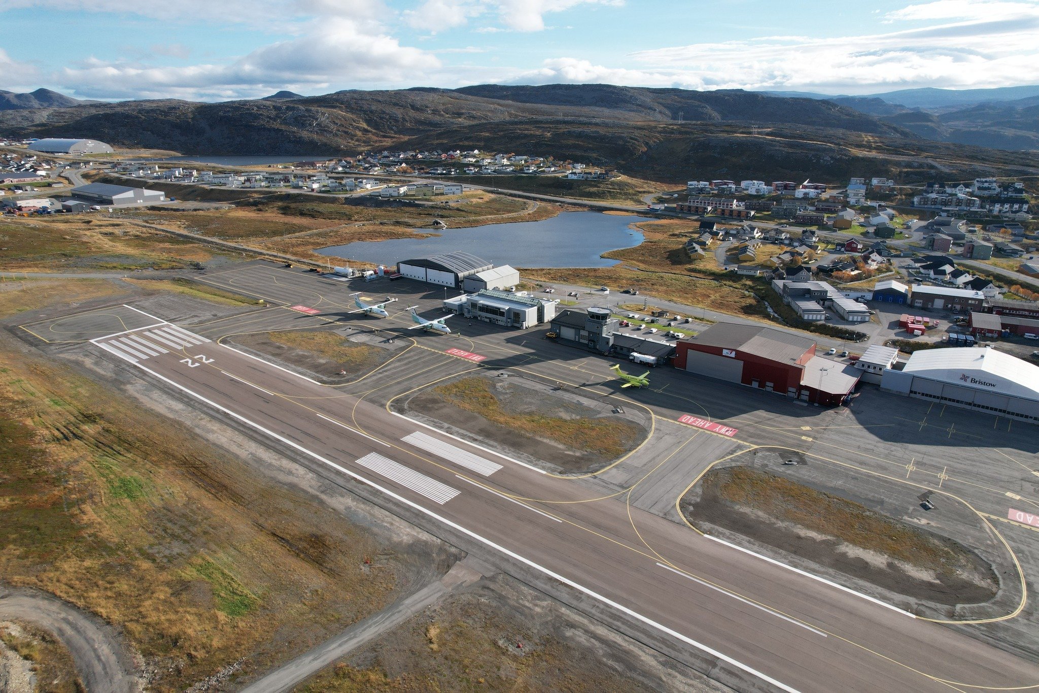 Flyfoto av Hammerfest flyplass med rullebane og terminalbygg omgitt av kupert landskap og en bygd i bakgrunnen.