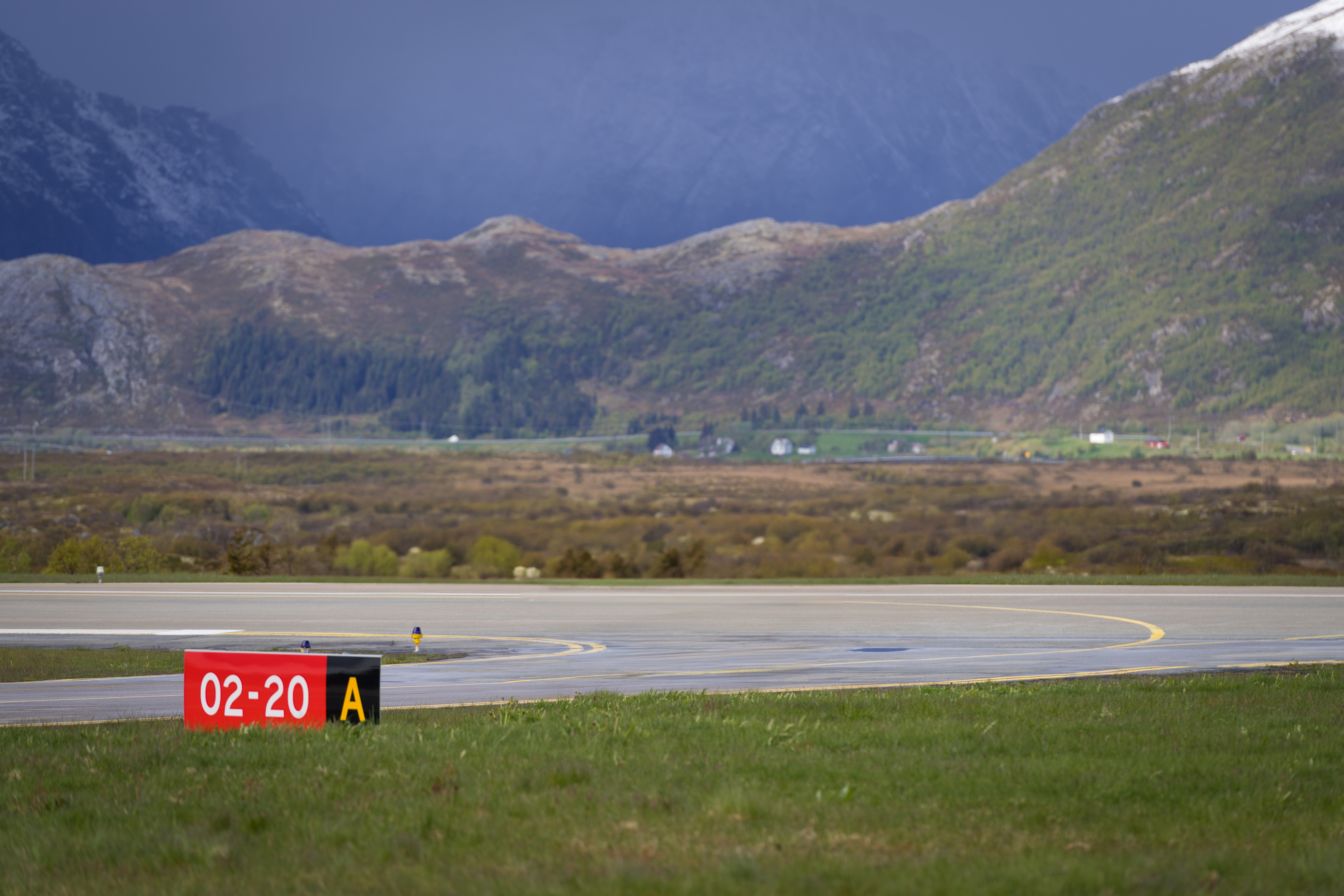 Flyplassrullebane med skilt 02-20 A, omgitt av norsk fjellandskap og grønne daler under overskyet himmel.