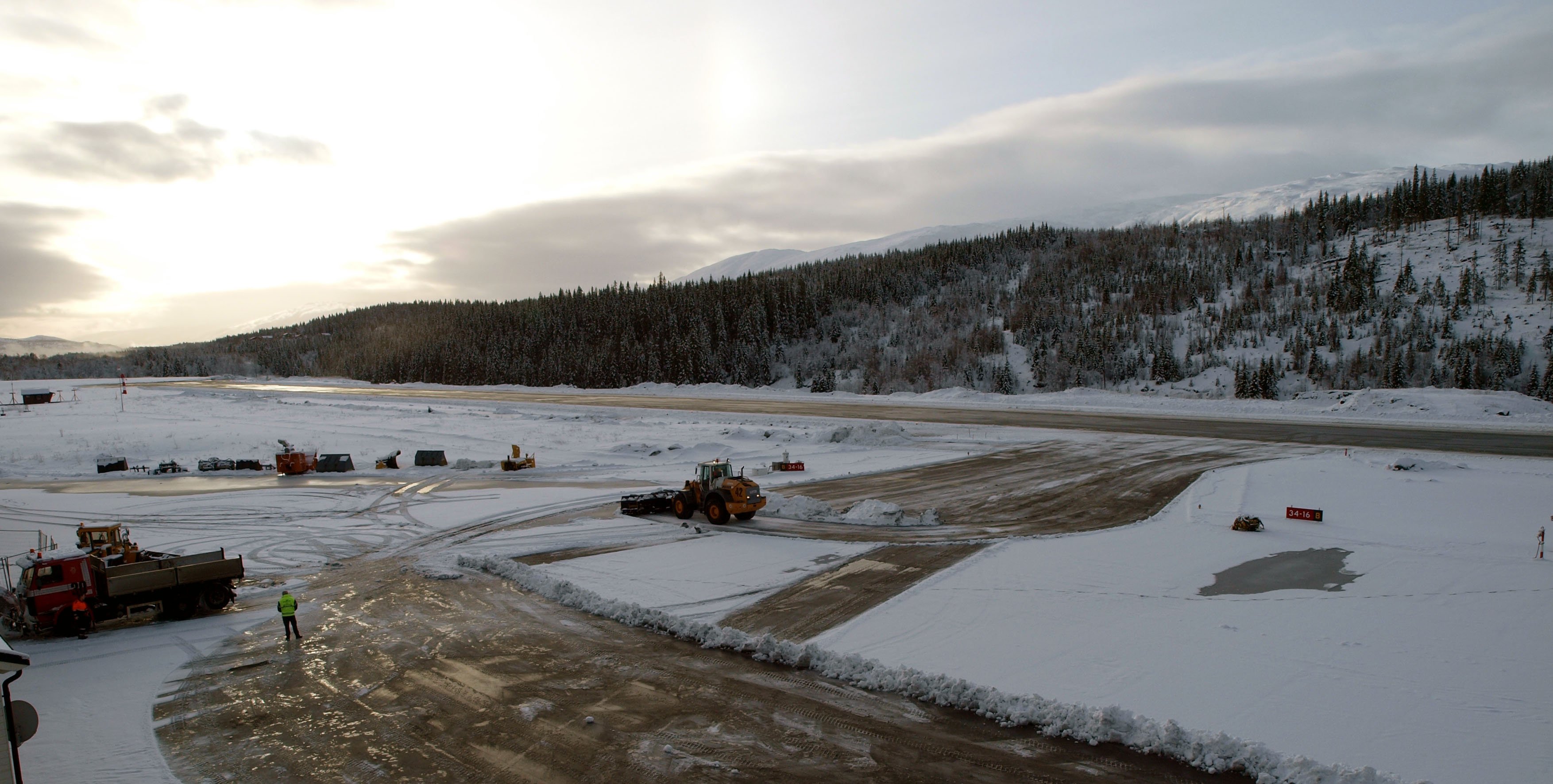 Snødekt flyplass med flystripe i vintersol, omgitt av skogkledde åser. Snøryddingsutstyr er i arbeid på plassen.