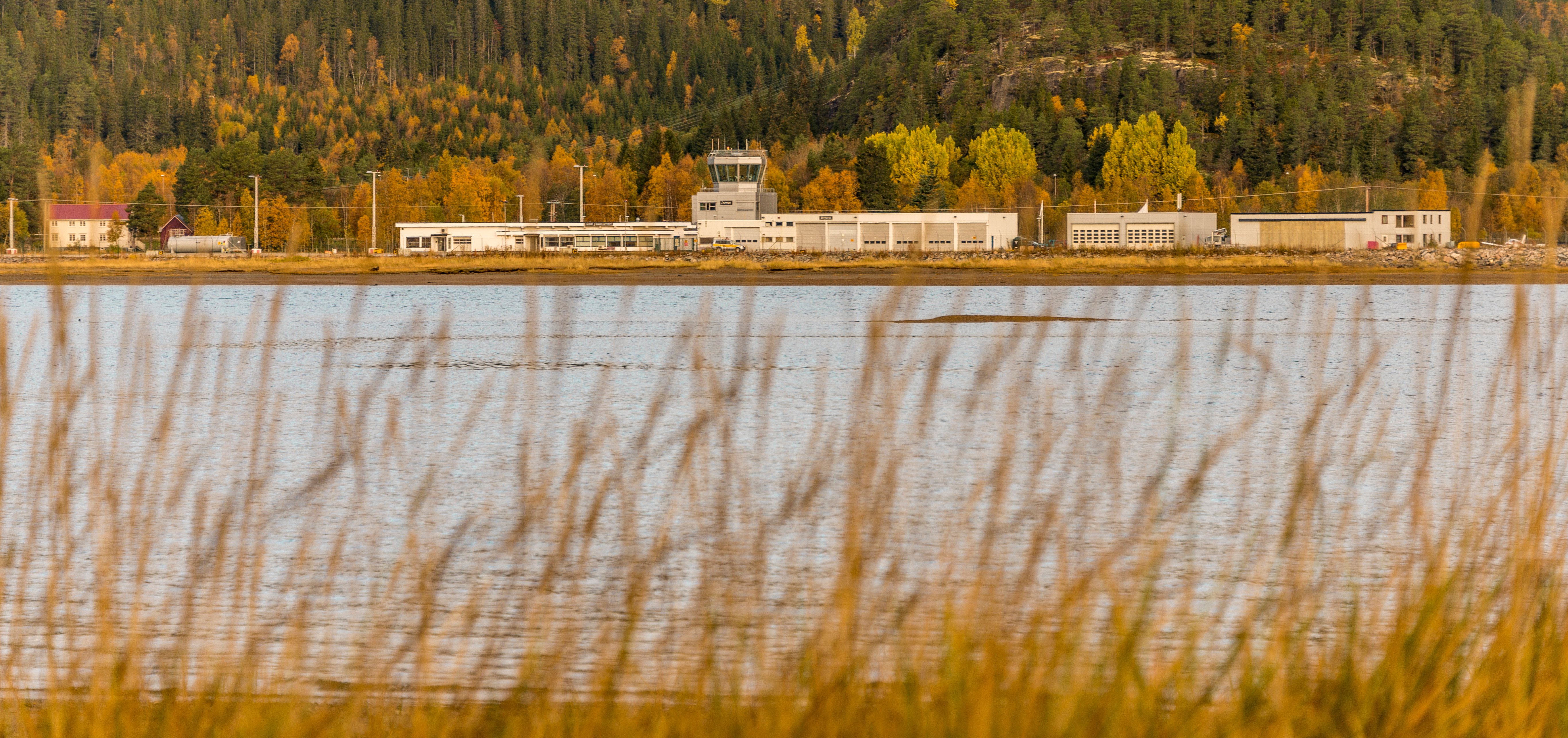 Flyplassbygning ved innsjø om høsten, omringet av høstfargede trær og åser i bakgrunnen, med siv i forgrunnen.