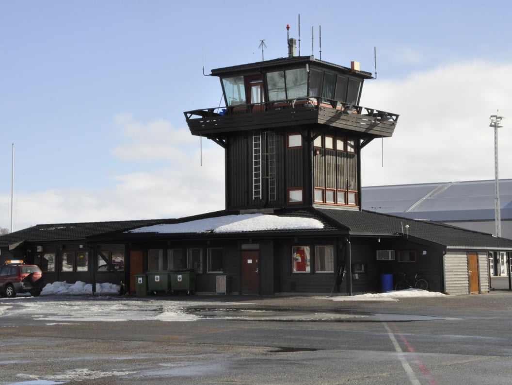 Control tower at Røros Airport with snow on the roof, a parking lot in front, and a car to the left. Winter landscape under a clear sky.