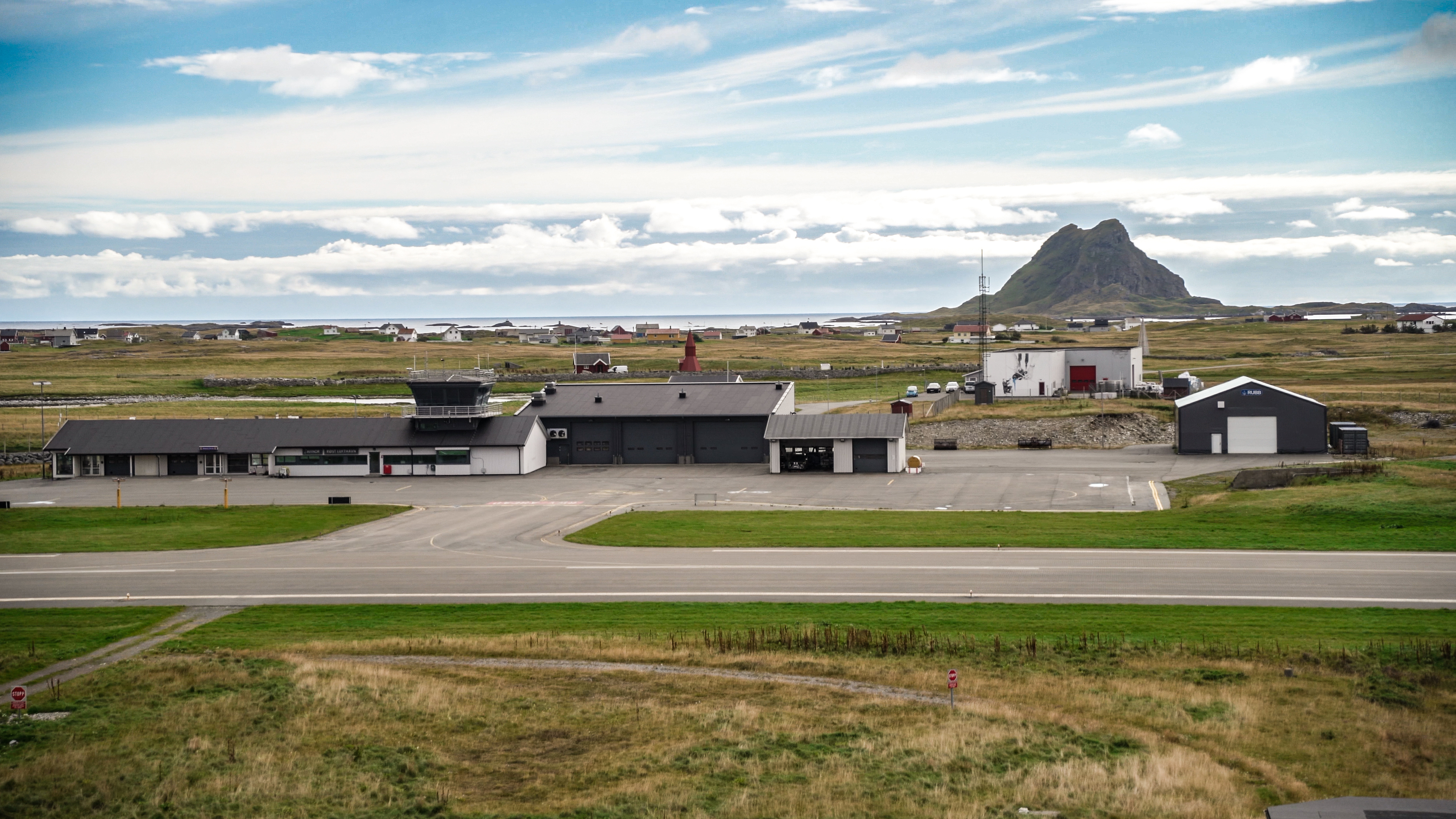 Flyplassbygning med landingsstripe i et landlig landskap med fjell i bakgrunnen under en delvis skyet himmel.