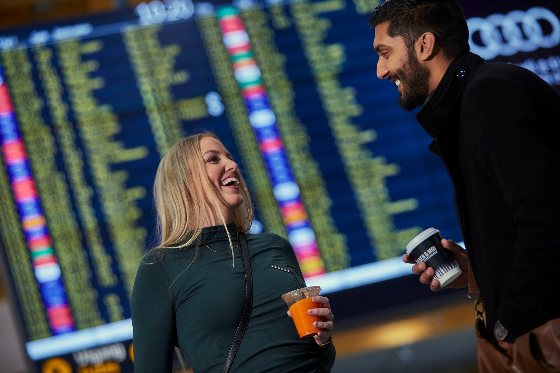 Two people laughing and holding drinks in front of a large airport departure board displaying flight information