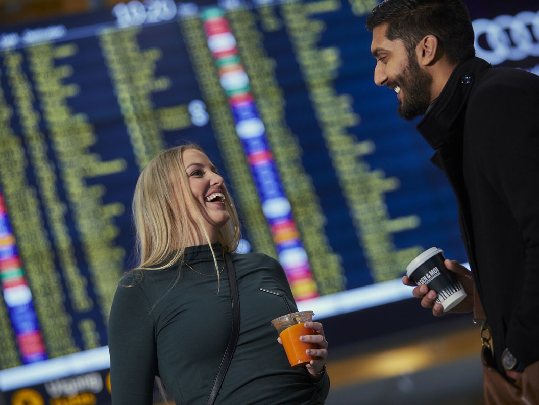 Two people laughing and holding drinks in front of a large airport departure board displaying flight information