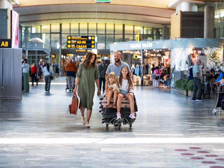 Family with luggage cart at the airport, among travel shops and departures.