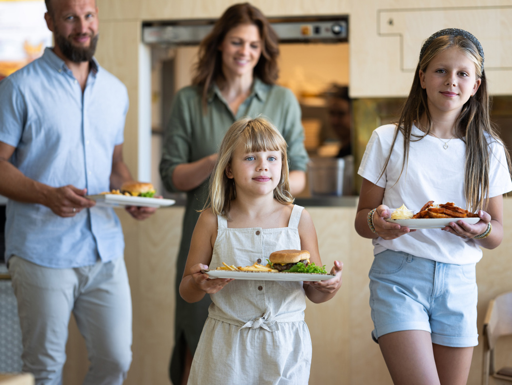 Familie med to barn som nyter et måltid på en restaurant, serverer hamburgere og pommes frites. Foreldrene smiler i bakgrunnen.