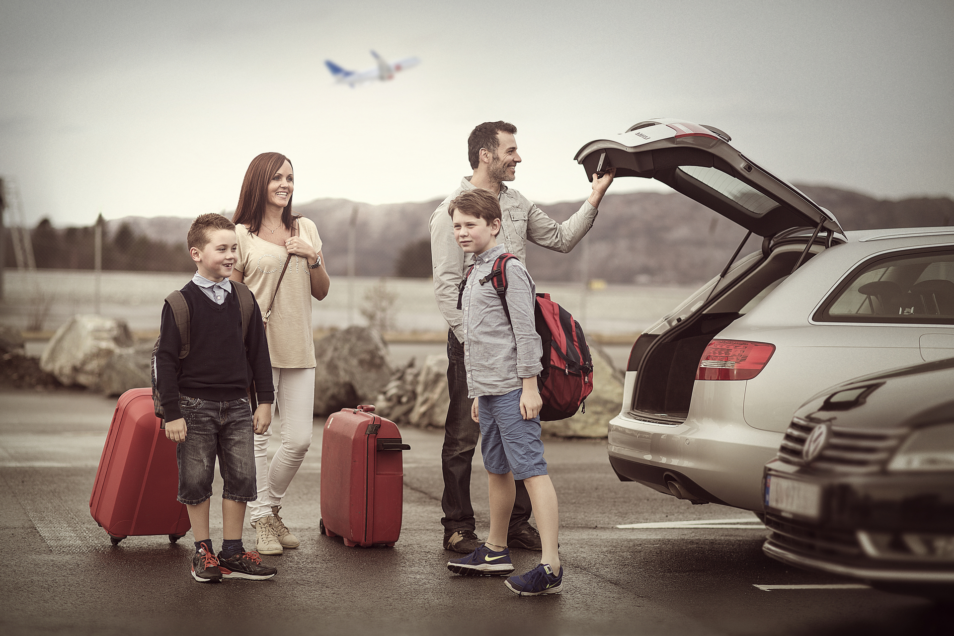 Familie på fire ved bil parkeringsplass, med kofferter og rygsekker klare for reise.