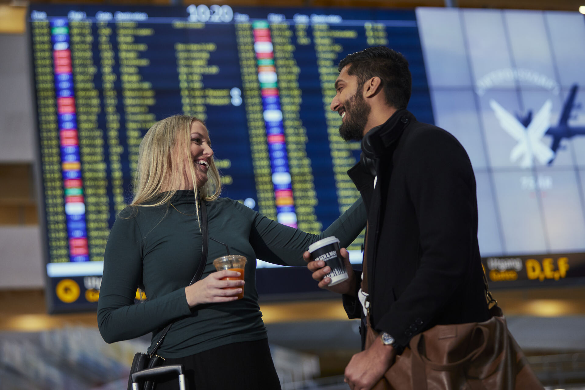 To personer på en flyplass smiler og holder drikke, i bakgrunnen vises en avgangstavle.
