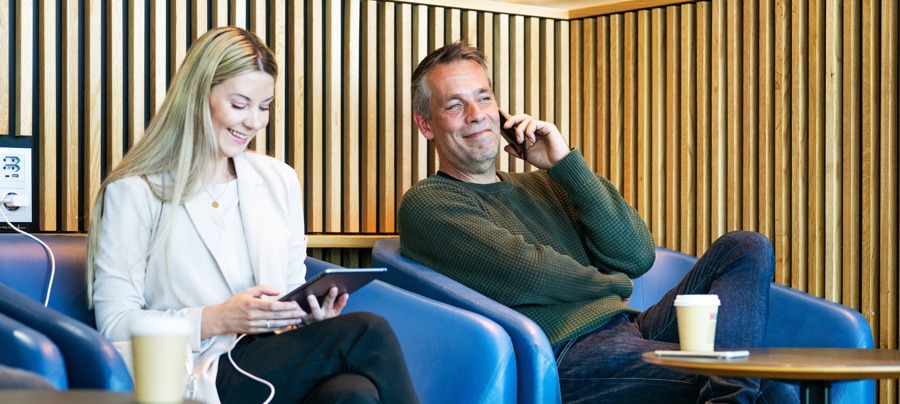 To personer slapper av i en moderne kafé. En kvinne ser på et nettbrett og en mann snakker i telefon. Begge sitter i blå stoler med kaffekopper på bordet foran seg.