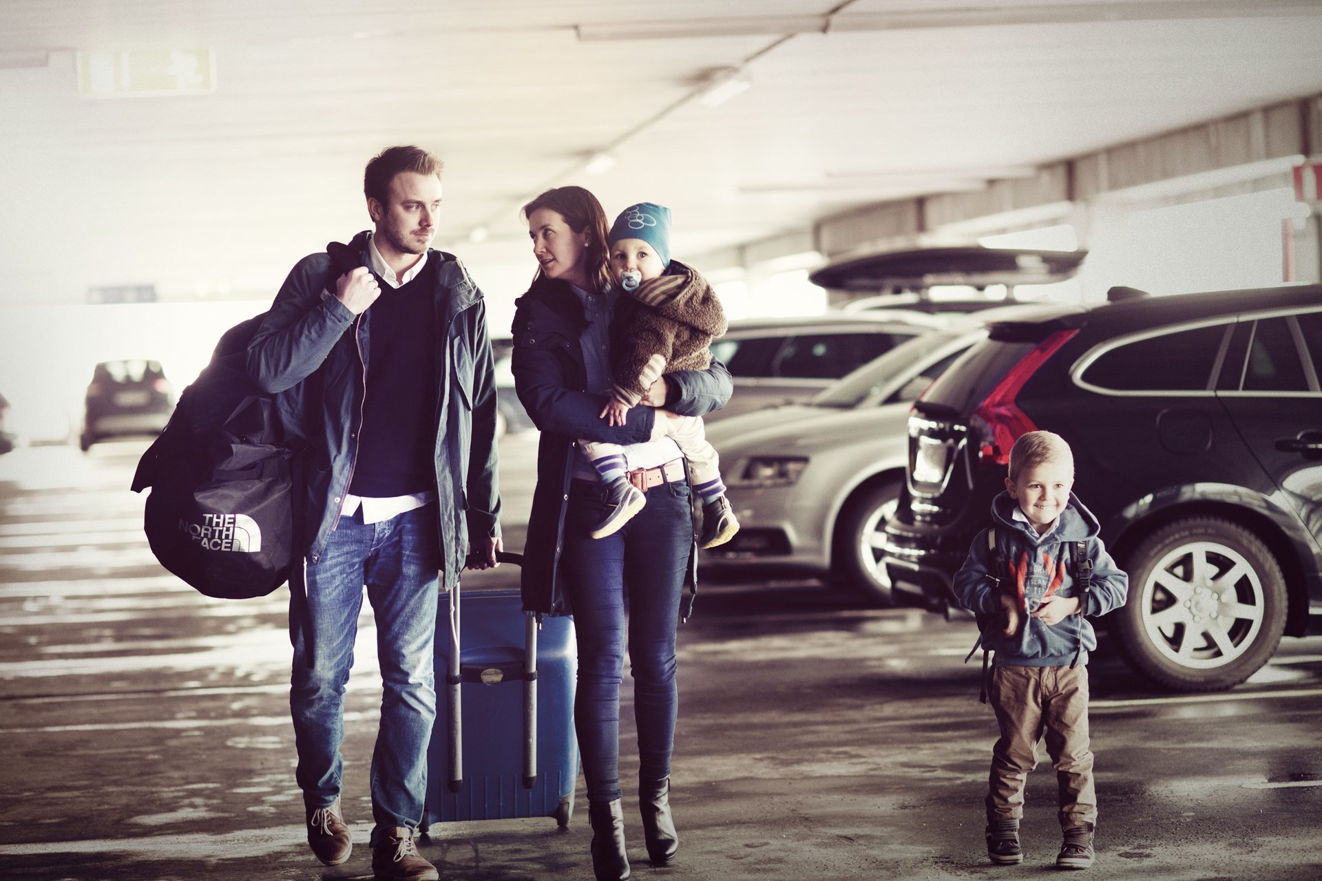Familie på fire med bagasje i parkeringshus, klare for reise. Foreldre og barn står blant biler med kofferter og ryggsekker.