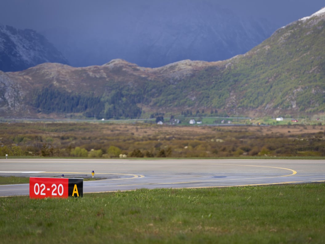 Airport runway with sign 02-20 A, surrounded by Norwegian mountain landscape and green valleys under an overcast sky.