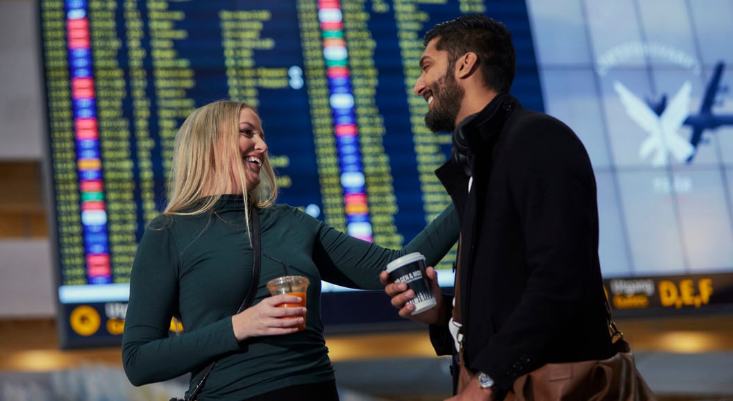 To personer på en flyplass smiler og holder drikke, i bakgrunnen vises en avgangstavle.