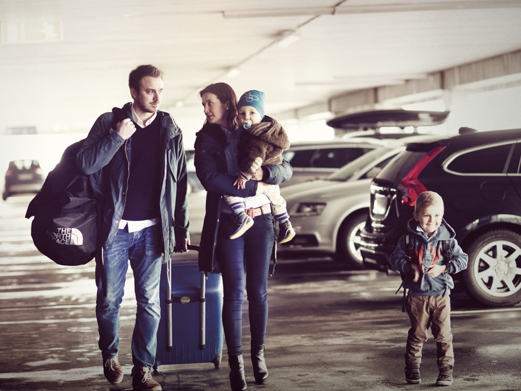 Familie på fire med bagasje i parkeringshus, klare for reise. Foreldre og barn står blant biler med kofferter og ryggsekker.