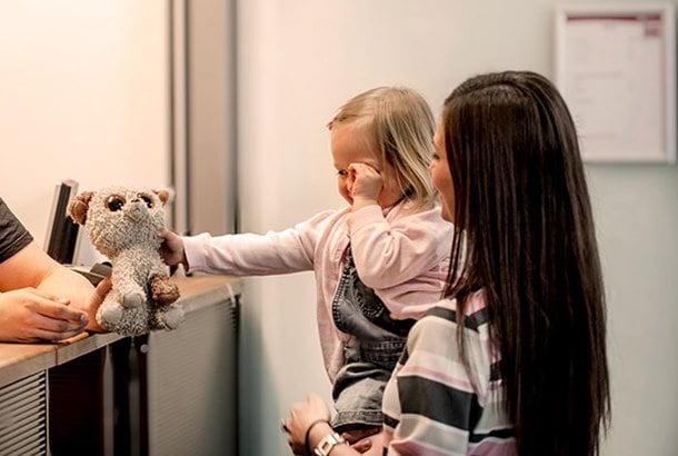 En dame med langt brunt hår står med en liten jente på armen foran en skranke hvor jenta får tilbake en bamse hun har glemt på flyet eller lignende.