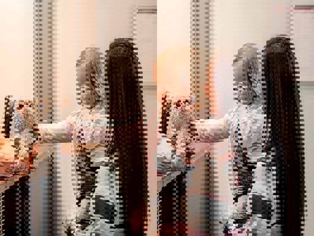 A lady with long brown hair stands with a little girl on her arm in front of a counter where the girl gets back a teddy bear she has forgotten on the plane or similar.