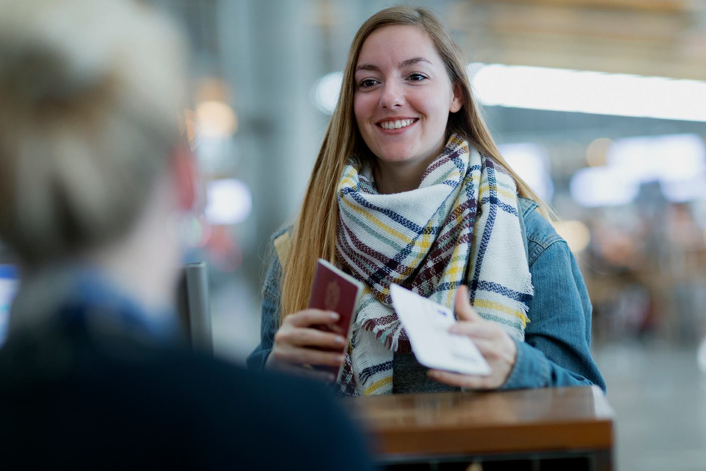 En dame med langt halvmørkt står foran en skranke på flyplassterminalen med passet sitt i hånden.