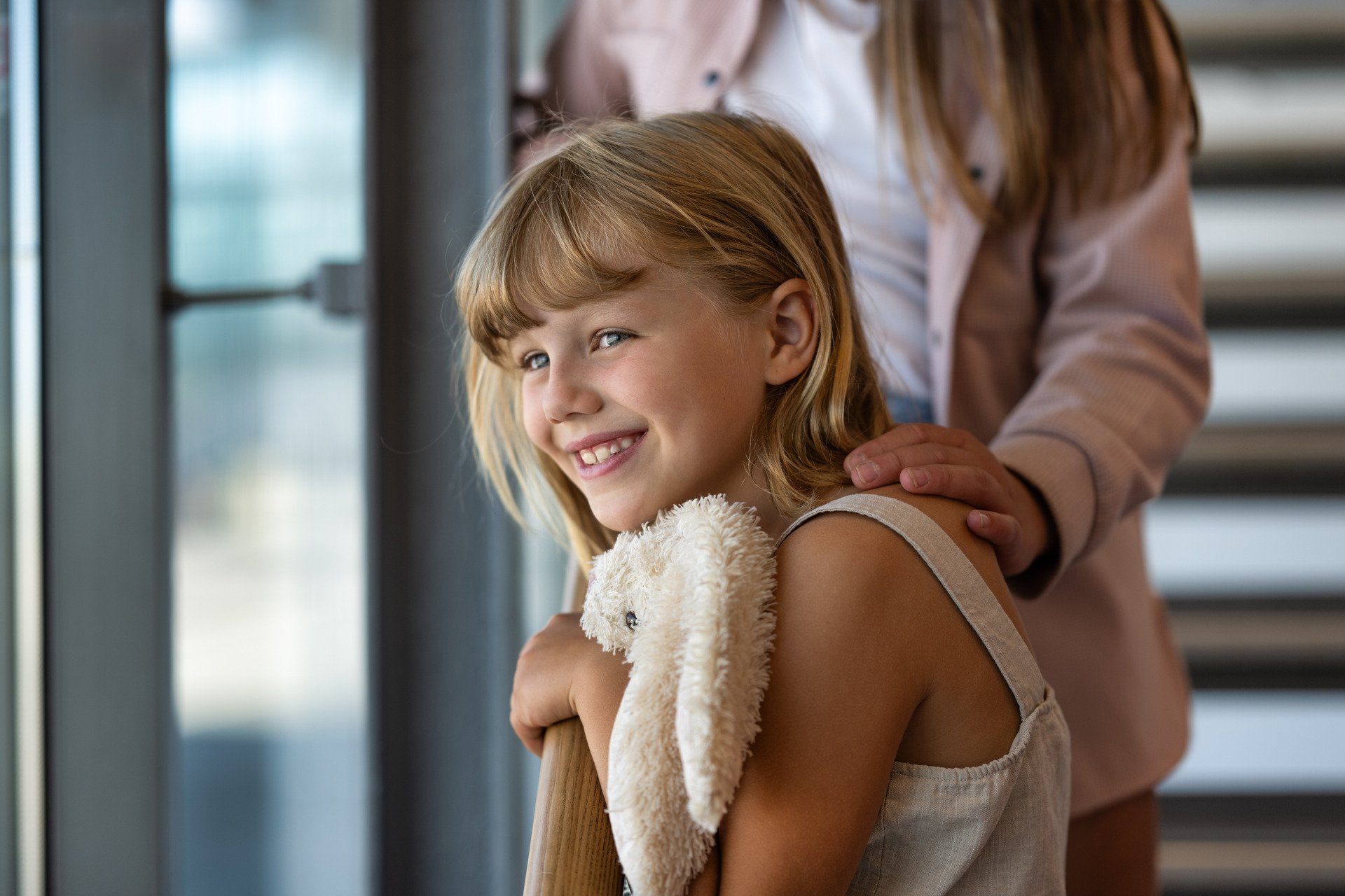 Jente med blondt hår smiler mens hun holder en myk bamse. En voksen persons hånd hviler forsiktig på skulderen hennes. Naturlig lys fra vindu i bakgrunnen.