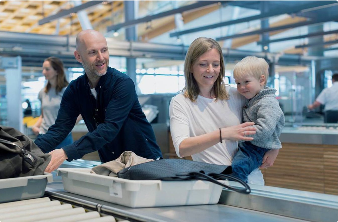 En mann og en dame som holder et lite barn står i sikkerhetskontrollen og legger tingene sine i grå esker som sendes avgårde. De er glade og smiler til hverandre.