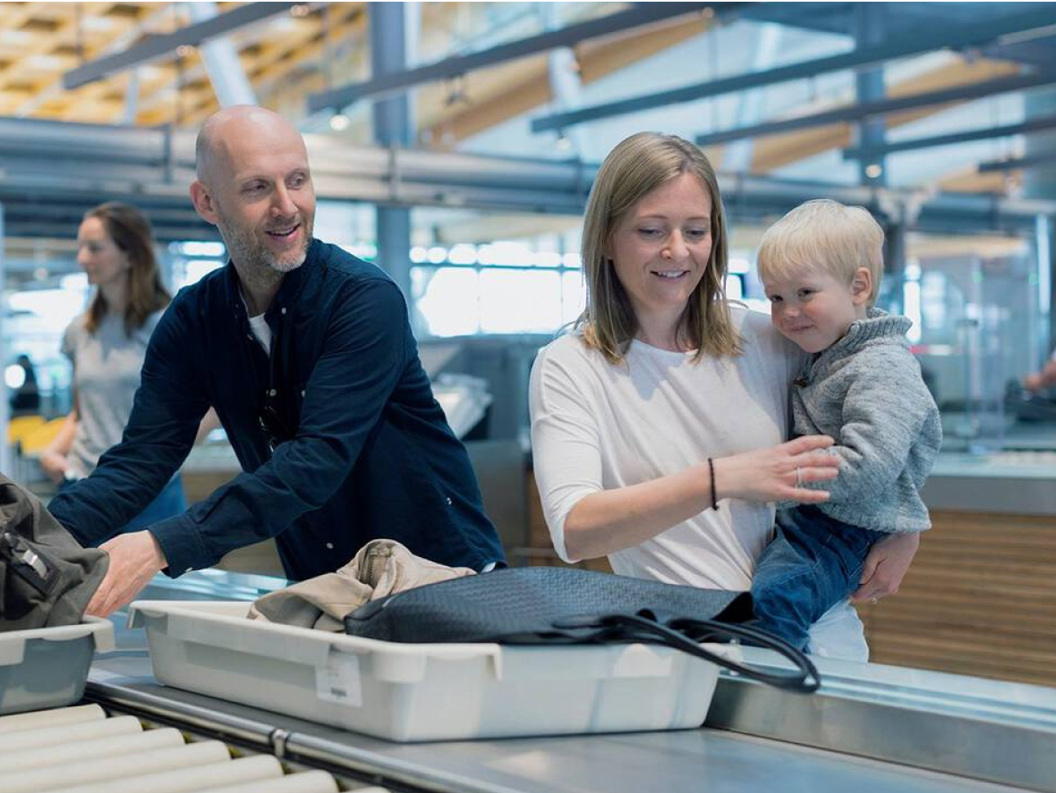 A man and a woman holding a small child stand at the security checkpoint, placing their belongings in gray bins that are sent off. They are happy and smile at each other.
