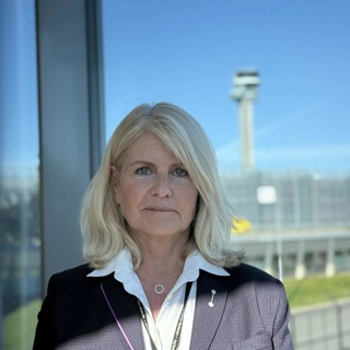 Woman in a blazer with a white shirt standing in front of a glass window overlooking an airport terminal and control tower in the background.