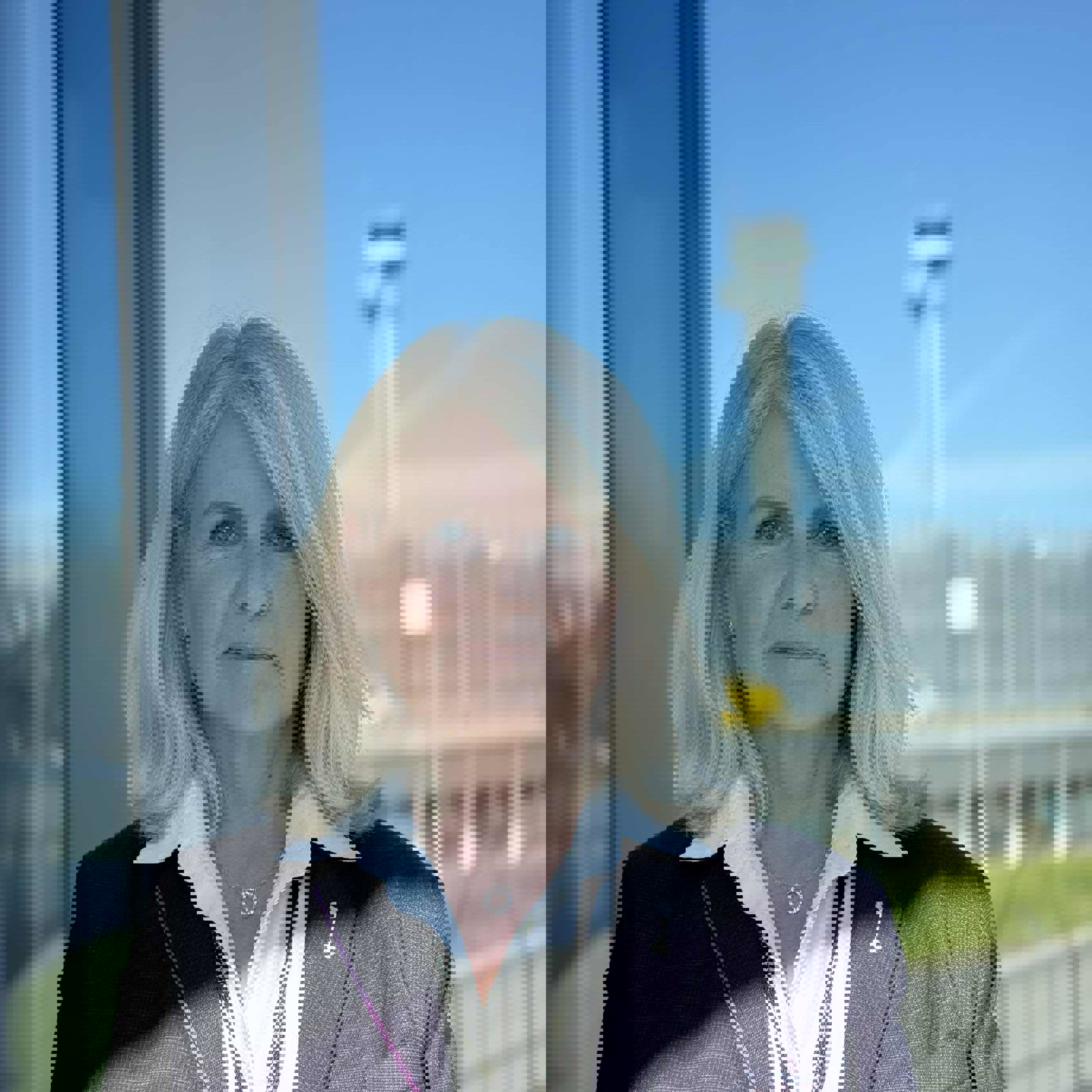 Woman in a blazer with a white shirt stands in front of a glass window overlooking an airport terminal and control tower in the background.